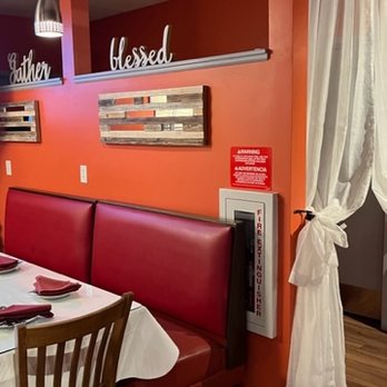 Bolly Twist - The image shows a section of a restaurant interior with a red upholstered booth seat along the wall, a wooden dining chair, and a table set with napkins and utensils. The wall behind the booth is painted in a warm red tone and decorated with a rectangular piece of wall art made of horizontal wooden slats. There are decorative words on a shelf above the wall art, including 'blessed.' To the right, a white curtain is tied back, revealing part of a doorway or window. The flooring is wood, and a fire safety panel is mounted on the wall near the curtain. - Indian Near Me - Stuart, Florida