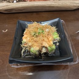 A sushi roll is presented on a black square plate, topped with crispy fried noodles and chopped green onions. The roll contains visible ingredients such as avocado and possibly seafood, with soy sauce underneath. The plate is placed on a wooden table, with a decorative tray and pinecones in the background.