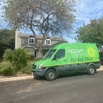 Ageint Security - Ageint Security Cameras Installation - A green security service van with the company name, logo, and contact number is parked on the street in front of a two-story residential house. The house has a gray exterior with white trim, multiple windows, and is surrounded by trees and shrubs. The sky is partly cloudy, and there is another vehicle parked nearby. - Home Automation Near Me - Conroe, Texas
