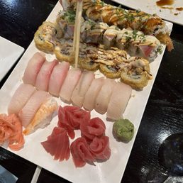 A rectangular white plate holds an assortment of sushi, including various sushi rolls topped with sauces and garnishes, slices of sashimi, and a small dollop of wasabi. The sushi rolls include different types with toppings like eel sauce, mayonnaise, and chopped green onions. Sashimi slices are arranged in a row, with pinkish fish and shrimp. The plate is placed on a dark wooden table, with small dishes of soy sauce visible in the background.