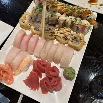 Sushi Naper - A rectangular white plate holds an assortment of sushi, including various sushi rolls topped with sauces and garnishes, slices of sashimi, and a small dollop of wasabi. The sushi rolls include different types with toppings like eel sauce, mayonnaise, and chopped green onions. Sashimi slices are arranged in a row, with pinkish fish and shrimp. The plate is placed on a dark wooden table, with small dishes of soy sauce visible in the background. - Sushi Bars Near Me - Naperville, Illinois