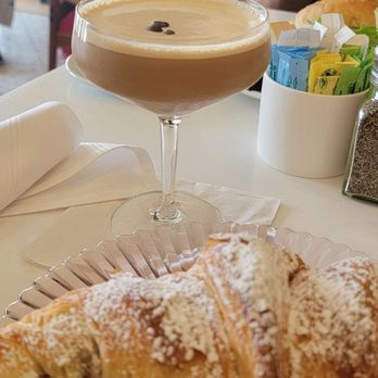 La Sorella - A pastry filled with strawberries and cream, dusted with powdered sugar, is on a decorative plate in the foreground. Behind it, a coffee cocktail with a frothy top and a few coffee beans is served in a coupe glass. The table has a napkin, a container with sugar packets, and a busy restaurant background with people dining and waitstaff serving. - Breakfast & Brunch Near Me - Fishkill, New York