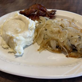 Pegah's - Liver and onions with bacon and extra onions, mashed potatoes - A plate featuring mashed potatoes with gravy, sautéed onions, and a strip of bacon, served on a white dish. - Salad Near Me - Lee's Summit, Missouri