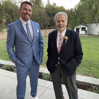 Savage Suiting - Dudes in suits - Two men in suits pose outdoors in a garden setting, smiling at the camera. One wears a light blue suit, the other a dark suit with a boutonniere. - Formal Wear Near Me - Placentia, California