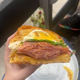 Chorizo Egg Sandwich - A breakfast sandwich with layers of ham, a fried egg, and fresh greens is held in a person's hand outdoors. The sandwich is made with toasted bread and appears freshly prepared. In the background, there is a white plate with some remaining food and an outdoor setting with furniture and plants, indicating a casual outdoor meal.