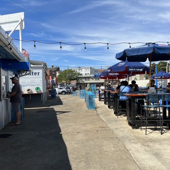 The Gloucester House - Outdoors - A seafood restaurant with outdoor seating under umbrellas, with patrons dining and a person ordering at a window, on a sunny day. - Seafood Near Me - Gloucester, Massachusetts