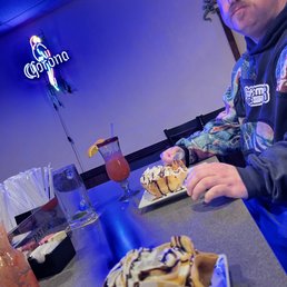 Deep fried ice creams and Korey - A man sits at a table with two plates of ice cream sundaes and two cocktails, in a restaurant with a neon Corona sign on the wall.