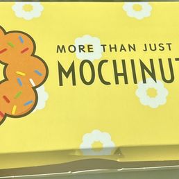 Box of 3 - The image shows a yellow box from Mochinut with a large donut illustration decorated with colorful sprinkles. The box has playful text that says 'More than just a donut' and includes small illustrations of a hot dog and a flower-shaped snack. The background of the box is decorated with white flower patterns, creating a cheerful and inviting appearance.