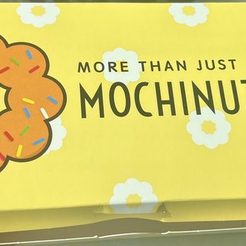 Mochinut-Gilbert - Box of 3 - The image shows a yellow box from Mochinut with a large donut illustration decorated with colorful sprinkles. The box has playful text that says 'More than just a donut' and includes small illustrations of a hot dog and a flower-shaped snack. The background of the box is decorated with white flower patterns, creating a cheerful and inviting appearance. - Bubble Tea Near Me - Gilbert, Arizona
