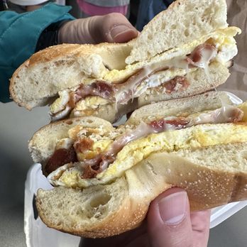 Cafe Fresh Bagel - Bacon and cheese breakfast bagel - A close-up of a bagel sandwich featuring scrambled eggs, melted cheese, and bacon, held by two hands. The bagel appears toasted and the filling is visible, with a background showing people in a casual setting. - Bagels Near Me - Dedham, Massachusetts