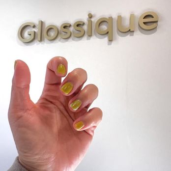 Glossique - A hand with yellow and glittery nail polish is positioned in front of a wall with the word "Glossique" in 3D letters. - Nail Salons Near Me - Pinole, California