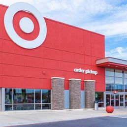 Target - The image shows the exterior of a Target retail store with a red building facade, a large white Target logo at the top left, and an area designated for order pickup. The storefront features large glass windows and a modern design with brick accents near the entrance. The sky above is partly cloudy, and the parking lot is visible in front of the store.