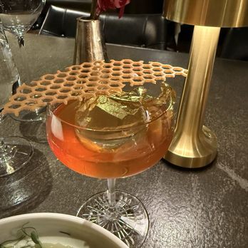 TOCA - Miele D'oro - A pinkish cocktail with ice cubes and a honeycomb-shaped garnish in a glass, placed on a restaurant table with a gold lamp and a vase with flowers. - Italian Near Me - Toronto, Ontario