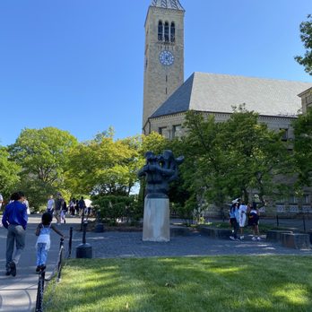 Cornell University - A historic church with a tall steeple and clock is set among green trees, with visitors walking nearby in a park-like area. - Colleges & Universities Near Me - Ithaca, New York
