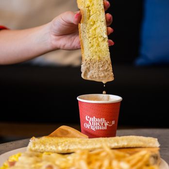 CAO Bakery & Cafe - A hand dipping a Cuban toast in Cuban coffee with milk. - Cuban Near Me - Fort Lauderdale, Florida CAO Bakery & Cafe - A hand dipping a Cuban toast in Cuban coffee with milk. - Cuban Near Me - Fort Lauderdale, Florida