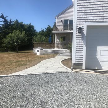 GrandScapes - a gravel driveway with a white garage door - A house with gray siding, a garage, and a curved concrete walkway leading to the backyard, which has trees and a deck. - Landscaping Near Me - Wakefield, Rhode Island