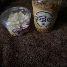 Mango pineapple with extra strawberries, extra bananas, and extra granola - Two acai bowls with various toppings are placed on a brown textured surface, featuring the logo of a cafe specializing in acai bowls and smoothies.