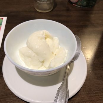 El Cedro - Mohavali - A bowl of vanilla ice cream is placed on a white plate with a metal spoon beside it, on a dark wooden table. - Lebanese Near Me - Brooklyn, New York