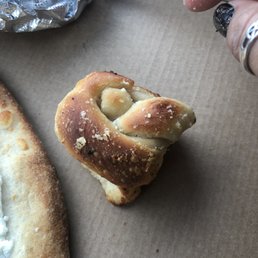 A piece of garlic bread with a toasted, golden-brown crust and visible garlic and herb bits rests on a brown paper surface. The bread appears soft inside with a slightly crispy exterior. Part of a pizza with cheese and a crust is visible on the left side of the image, and aluminum foil is seen in the top left corner. A person's finger with black nail polish and a silver ring is near the top right corner.