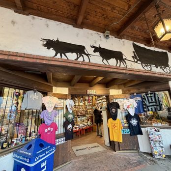 Covered Wagon - The image shows the entrance of a gift shop with clothing items such as T-shirts and sweatshirts displayed outside on hangers and mannequins. The shop has a rustic exterior with a wooden overhang and a mural of oxen pulling a covered wagon painted on the wall above the entrance. A blue recycling bin is placed near the entrance, and the shop's interior is visible through the open doorway, with more items on display inside. - Jewelry Near Me - Albuquerque, New Mexico