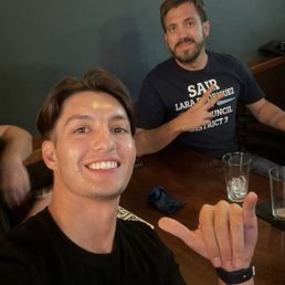 Sharing a drink and knocking out trivia questions with my friend Sair - Two men are seated at a wooden table in an indoor space with dark walls, one taking a selfie and the other making a peace sign.