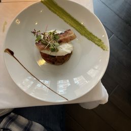 Octopus - A gourmet dish featuring microgreens, a slice of bacon, and a dollop of cream on a bed of grains, garnished with green sauce and a line of balsamic reduction.