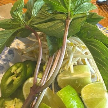 Pho Duy - Mint, bean sprouts, and lime - The image shows a plate with fresh herbs, including basil, lime wedges, bean sprouts, and sliced green peppers. The herbs are placed on a decorative plate, with a wooden table and additional dishes visible in the background. The ingredients are typical accompaniments for Vietnamese dishes like pho, emphasizing freshness and traditional flavors. - Vietnamese Near Me - Greeley, Colorado