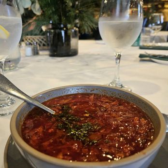 Noroc Restaurant - Borscht - A bowl of borscht, a traditional Eastern European beet soup, is placed on a restaurant table. The soup is garnished with fresh herbs, possibly dill. The table also has glasses of water, one with a lemon slice, and a decorative plant in the background. The setting appears to be a dining area with a white tablecloth and ambient lighting. - Desserts Near Me - Carmichael, California
