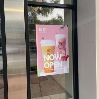Moge Tee - Sign on window - The image shows a glass storefront window with a pink poster advertising the opening of Moge Tee. The poster displays two beverage images, one with a yellowish foam tea and the other with a pinkish bubble tea, along with the text 'NOW OPEN' and additional details about the drinks. The window reflects trees and the sky outside, and the building has a modern exterior with a stone or concrete wall. - Bubble Tea Near Me - Lynnfield, Massachusetts
