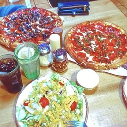 small 12 inch pepperoni hot green chili pizza and a house dinner salad with ranch dressing. - The image shows a wooden table with two large pizzas, one topped with pepperoni and green chili, and the other with pepperoni and basil. A plate of salad with cherry tomatoes, lettuce, and shredded cheese is also present, along with two glasses of soda and a glass of water. Condiment shakers and a small bowl are on the table, and a black chair is visible in the background, indicating a casual restaurant environment.