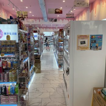 Shibuyala - The image shows a store aisle with white marble-like tiled flooring and shelves on both sides stocked with snacks, toys, and miscellaneous products. The ceiling is painted pink with a decorative drip pattern and has multiple ceiling lights and a large blue duct running across. A person is visible at the far end of the aisle, shopping with a pink basket. Promotional signs and advertisements are hanging from the ceiling and attached to the shelves, indicating various products and offers. - Cosmetics & Beauty Supply Near Me - Boston, Massachusetts