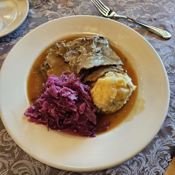 Garden Grill - Sourbratten - A white round plate holds slices of roast beef covered in gravy, accompanied by a serving of mashed potatoes and cooked red cabbage. The plate is placed on a patterned tablecloth, with a fork and other tableware visible nearby, indicating a meal setting at a restaurant or home dining table. - Seafood Near Me - Smithtown, New York