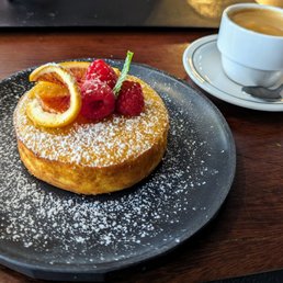 Clementine Cake - A round dessert, possibly a cake or French toast, is presented on a black plate, topped with fresh raspberries, orange slices, and a small green decoration, all dusted with powdered sugar. To the right, there is a white cup filled with coffee or espresso on a matching saucer with a spoon. The setting appears to be in a cozy café or restaurant with a wooden table surface.