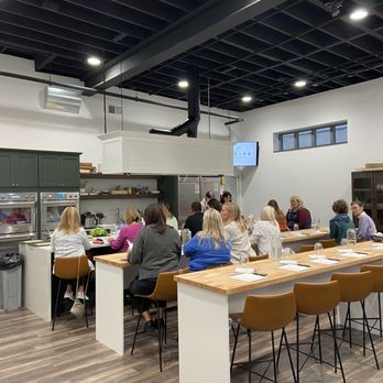 The Recipe - Class was full tonight - Participants are seated at a large kitchen island in a cooking class setting, with kitchen appliances and shelves in the background. - Venues & Event Spaces Near Me - Ames, Iowa