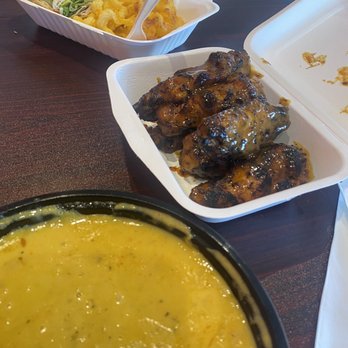 Chef BobbyD Restaurant & Catering - Such delicious food (barbecue wings, bacon wrap and lobster bisque)! - The image shows three takeout containers on a dark wooden table. One container holds grilled chicken wings coated in sauce, another contains a fresh salad with lettuce, purple onions, and other vegetables, and a third bowl contains a yellow soup with visible herbs and spices. The containers are open, revealing the food inside, and the setting appears to be a casual dining or takeout environment. - Caribbean Near Me - Baltimore, Maryland