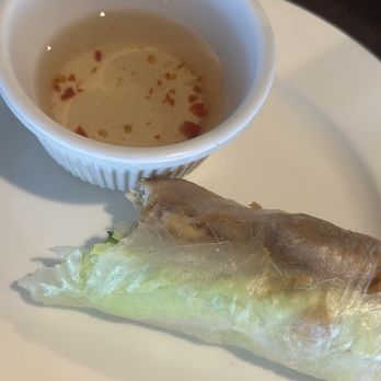 Pho Yummy - A fresh Vietnamese spring roll wrapped in rice paper, served with a small cup of dipping sauce containing chili flakes, on a white plate. - Vietnamese Near Me - Stuart, Florida