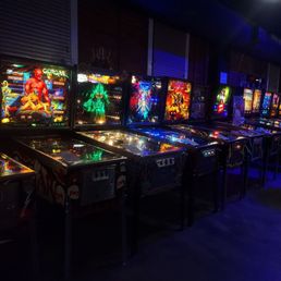Roughly 10 pinball machines lined up against a wall with dim purple lighting Roughly 10 pinball machines lined up against a wall with dim purple lighting
