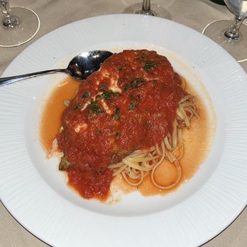 Camille's - Pollo Alla Parmigiano - A plate of chicken Parmesan with melted cheese and tomato sauce is placed on a white dinner plate, served over a bed of spaghetti. The dish is garnished with chopped herbs. The table setting includes a spoon, wine glasses, and beverages, indicating a restaurant dining environment. - Italian Near Me - Providence, Rhode Island