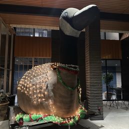 Goose outside of lobby - A large goose sculpture, positioned indoors, is decorated with string lights, tinsel, and Christmas ornaments, including a green garland around its neck. The sculpture is placed on a small trailer with a Colorado license plate, and the background features windows, a brick column, and indoor furniture. The setting appears to be a commercial or public space with holiday decorations.