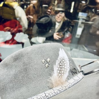 Kemo Sabe - creating my hat- simple monochrome grey vibes - A grey felt hat featuring a feather and decorative band is positioned behind a glass window. The reflection in the glass shows several people, some holding phones, and the interior of a store with various items on display. The hat has a textured band with a pattern resembling scales and metallic embellishments. The image captures both the hat and the reflections of shoppers and store environment. - Shoe Stores Near Me - Aspen, Colorado