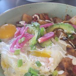 A breakfast plate featuring fried eggs with runny yolks, sliced avocado, pickled red onions, chopped green onions, and diced potatoes.