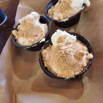 Mission BBQ - Three small black cups of ice cream are arranged on a brown paper-lined tray. Each cup contains a scoop of ice cream topped with cookie crumbs, with some crumbs scattered on the surface. The background shows a wooden table and part of a drink, indicating a casual dining environment. - Barbeque Near Me - Springfield, Illinois