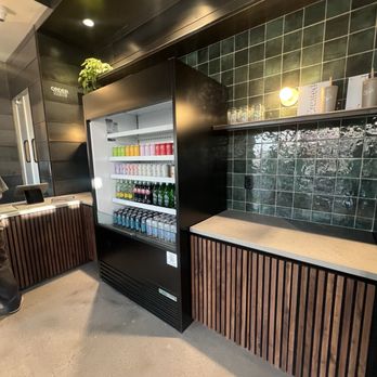CoffeeDrunk - A black beverage refrigerator filled with bottled drinks is placed against a dark green tiled wall in a contemporary setting. - Coffee & Tea Near Me - Phoenix, Arizona