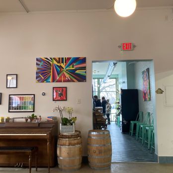 Cloud Walking Coffee - The image shows the interior of a coffee shop with white walls decorated with colorful artwork, including abstract and animal-themed pieces. A wooden piano is positioned against the wall, with small decorative items and books on top. Two wooden barrels are placed in front of the piano, one with a potted plant. To the right, an open doorway leads to a seating area with large windows, where customers are gathered. The space has a mix of modern and rustic decor, with green chairs stacked along the wall and natural light streaming in. - Chocolatiers & Shops Near Me - South Bend, Indiana