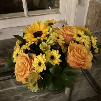OpenBlooms - A bouquet of mixed flowers, including yellow sunflowers, peach roses, and yellow daisies, is arranged in a glass container on a glass table. The background features a window with a white frame and a brick wall, with some objects visible through the window. The setting appears to be indoors, near a rustic brick wall and a window, with the flowers as the focal point. - Florists Near Me - Chantilly, Virginia
