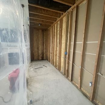 GarBros Builders - The image depicts an interior construction site with exposed wooden wall studs, electrical wiring, and unfinished ceiling joists. The floor is bare concrete, and there is a plastic sheet on the left side, possibly for dust protection. The space appears to be in the early stages of interior renovation or remodeling, with visible electrical outlets and wiring ready for further installation. - General Contractors Near Me