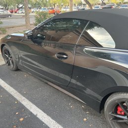 A black BMW convertible with a soft top is parked in a marked parking space, reflecting nearby trees and buildings on its surface.
