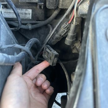 Firestone Complete Auto Care - On the end of the piece it says FUSE - The image shows a person's hand with dirt and grime on the fingers, pointing at a disconnected black electrical connector within a vehicle engine bay. The surrounding area includes various engine wires, hoses, and components, indicating maintenance or repair work. The engine bay appears dusty and used, with visible wiring and parts typical of automotive engine compartments. - Auto Repair Near Me - Oak Park, Illinois