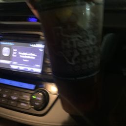 A plastic cup of iced tea is held in front of a car's dashboard, with the car's infotainment screen and controls visible in the background during nighttime.
