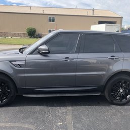 A gray Range Rover SUV with tinted windows and black wheels is parked in a parking lot near a beige industrial building with a small shrub.